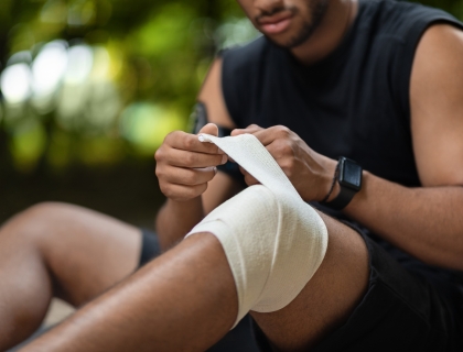 Unrecognizable african american sportsman sitting on the ground, wrapping injured knee with elastic bandage, got trauma while exercising outdoors. Injuries during physical activity concept